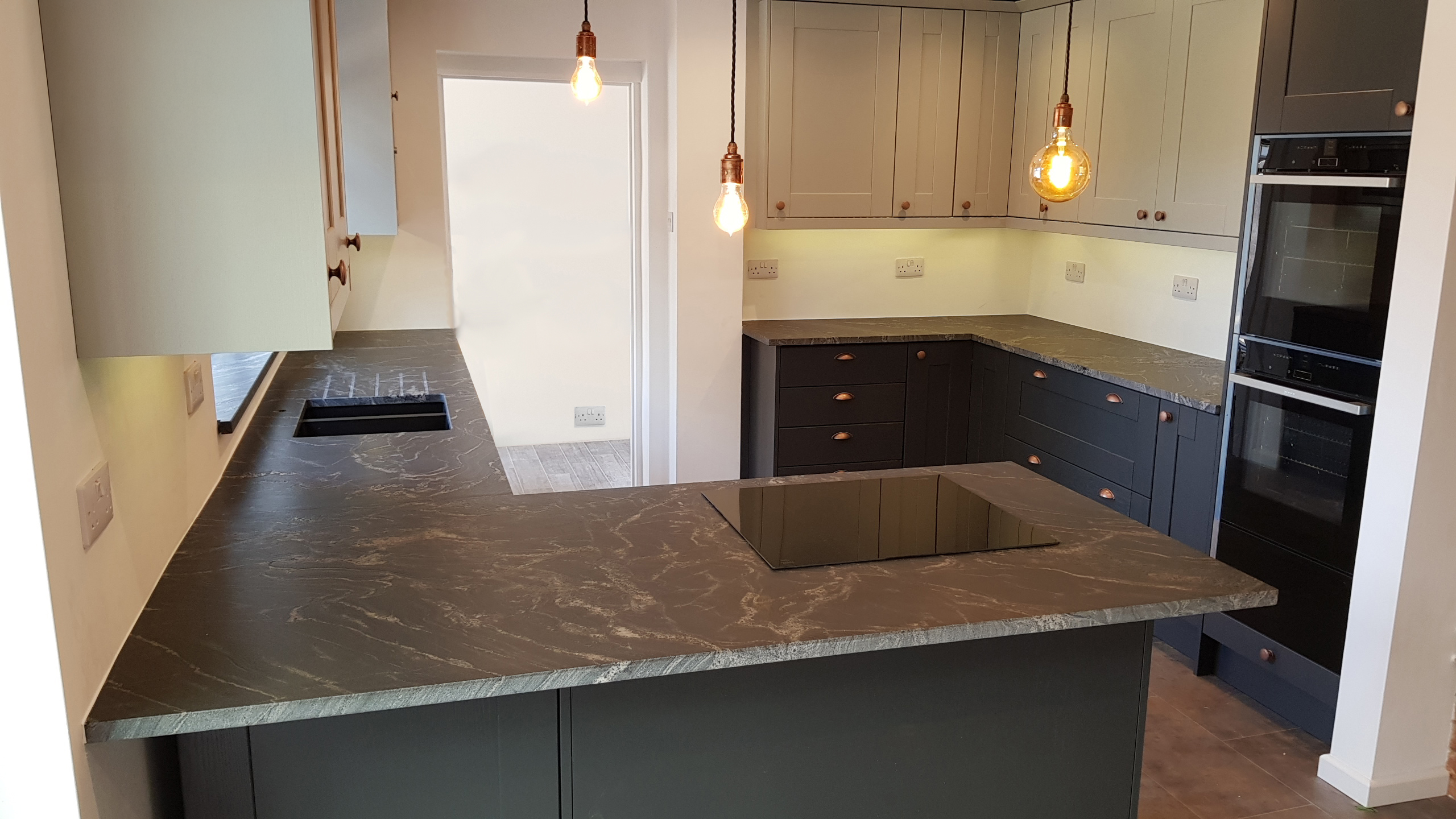 Wide angle view of a modern kitchen with Black Storm leathered Granite worktops on a peninsula and back run, showing the sink cut-out, hob area and pendant lighting.