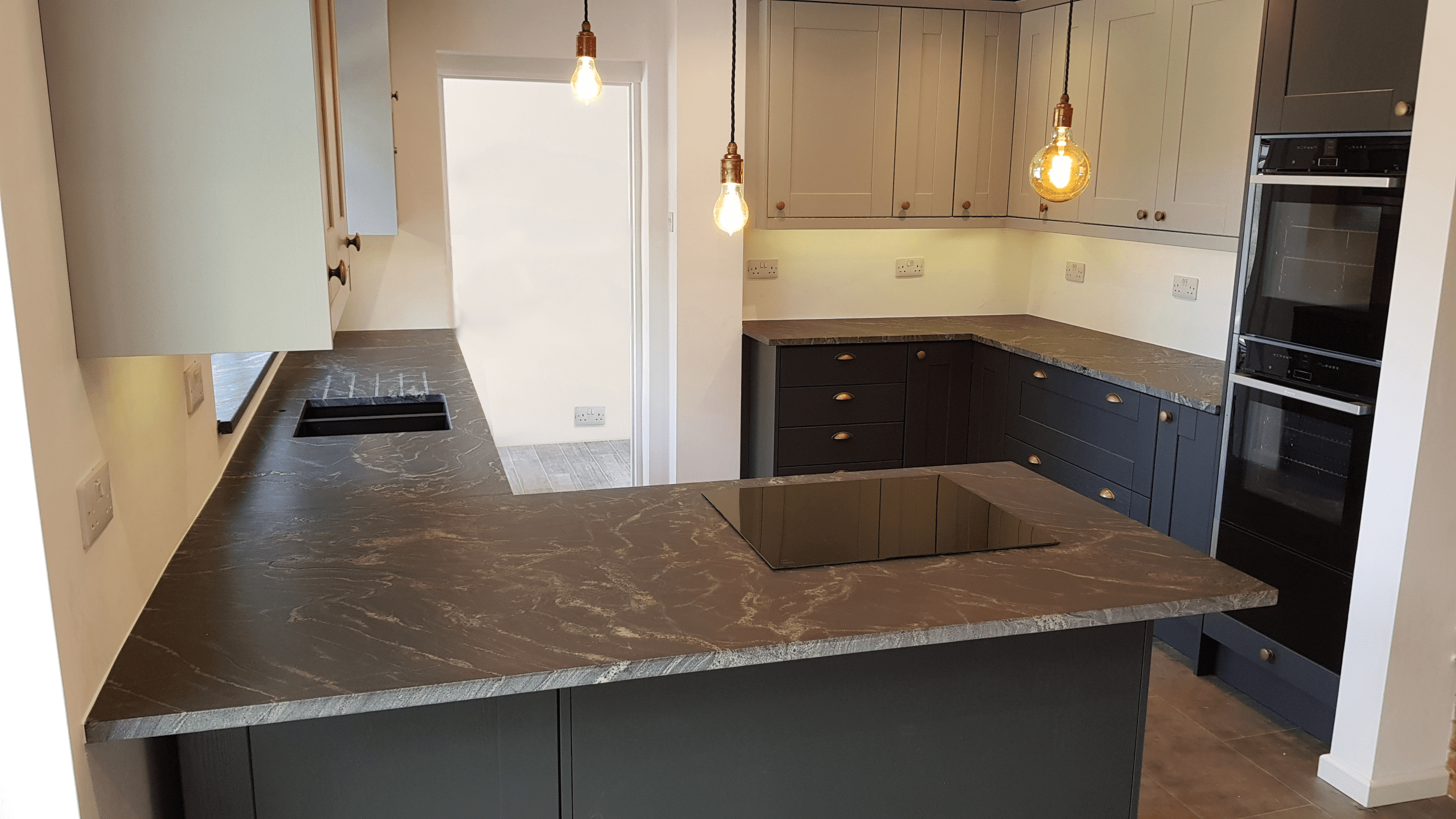 Wide angle view of a modern kitchen with Black Storm leathered Granite worktops on a peninsula and back run, showing the sink cut-out, hob area and pendant lighting.