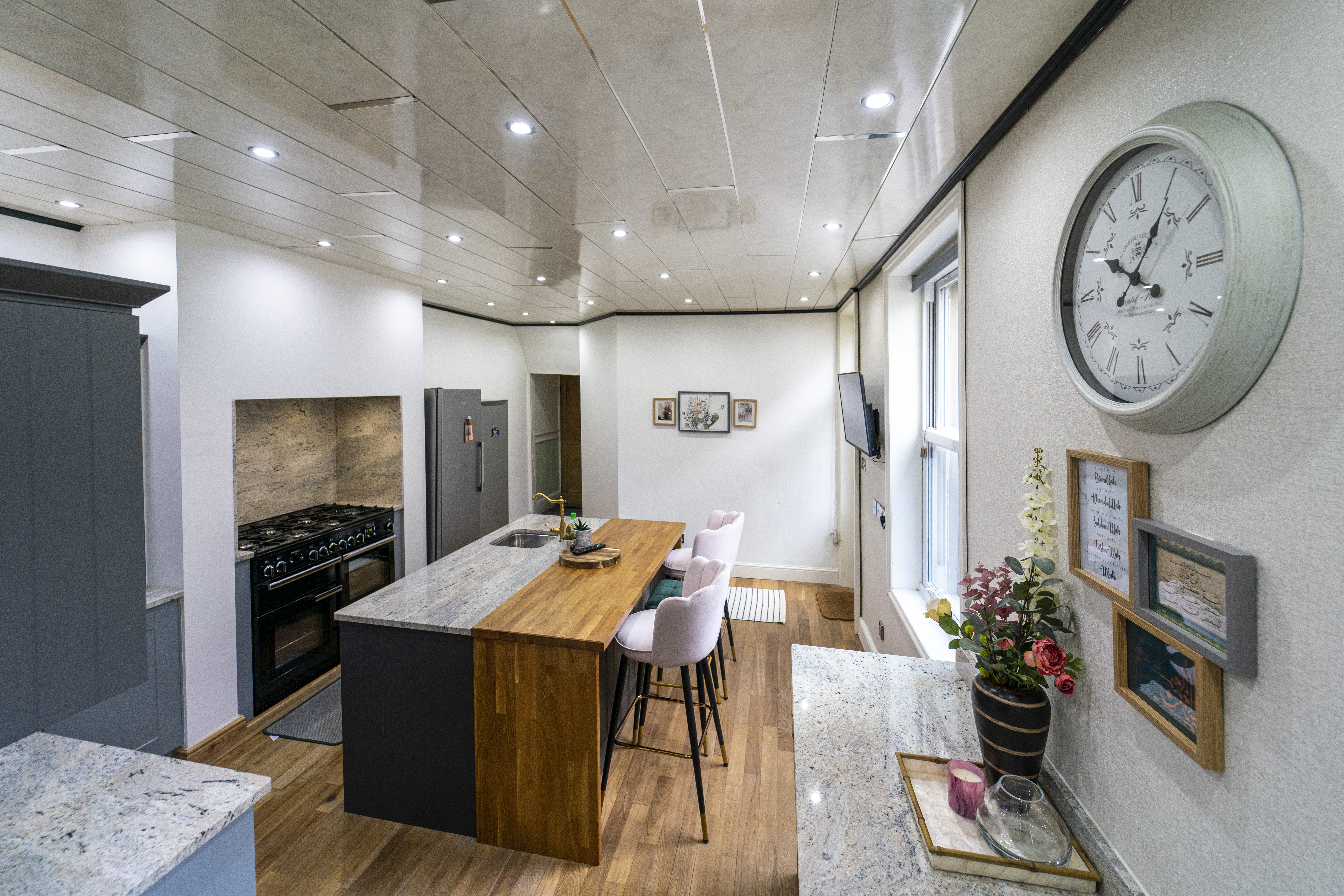 Wide angle view of a kitchen island with Ivory Fantasy Granite worktop and timber breakfast bar end, showing seating, range cooker and skylight ceiling.